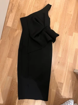 Svart cocktail midi klänning Asos Design strl 38 - Svart midi klänning från Asos Design i storlek 38. One shoulder, volang framtill och dragkedja i sidan.  Tyget är scuba tyg.   I nyskick, endast använd vid ett tillfälle. 
