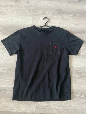 Svart t-shirt från Polo Ralph Lauren - Svart t-shirt från Polo Ralph Lauren med klassisk passform. T-shirten har en bröstficka med röd broderad logga och är tillverkad i 100% bomull. Perfekt basic-plagg med rund hals och korta ärmar. Skicket är Mycket bra nästan aldrig använd 100% äkta Storlek S