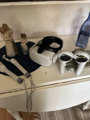 Oculus Quest 2 VR-headset med kontroller - Säljer ett Oculus Quest 2 VR-headset i vit plast med tillhörande två vita handkontroller. Headsetet har en svart justerbar rem och ergonomisk design för bekväm användning. Perfekt för gaming och VR-upplevelser hemma. En av kontrollerna har en spricka i sig men fungerar som vanligt ändå.