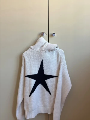 Vit stickad tröja med svart stjärna - Vit stickad tröja med en stor svart stjärna på ryggen. Tröjan har avslappnad passform, ribbstickade muddar och rund halsringning. Perfekt för dig som gillar en enkel men ändå cool look.