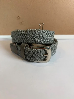 Grått flätat bälte med silverdetaljer - Snyggt grått flätat bälte med silvrigt spänne och detaljer i skinnimitation. Perfekt för att ge din outfit en clean och stilren touch. Bältet är flexibelt och passar till många olika looks.