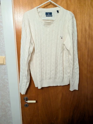 Vit kabelstickad tröja från Gant - Kabelstickad tröja i offwhite från Gant med rund halsringning och diskret logga broderad på bröstet. Tillverkad i mjuk premiumbomull och har ribbade muddar vid ärmslut och nederkant. Perfekt för en stilren och klassisk look.