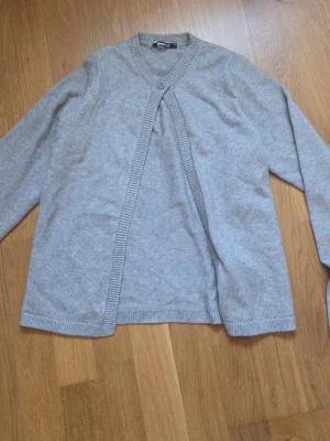 Ljusgrå kofta från Gina Tricot - Ljusgrå kofta från Gina Tricot med lång ärm och ribbade kanter. Koftan har en enkel knapp framtill vid halsen och är öppen nedtill. Perfekt att slänga på sig över en t-shirt eller linne för en chill look.