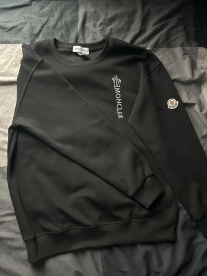 Svart sweatshirt från Moncler - Svart sweatshirt från Moncler med vit logotyptryck på bröstet och Moncler-märke på ena ärmen. Tröjan har rund halsringning, ribbade muddar och är tillverkad i mjukt material. Perfekt för en stilren och avslappnad look.
