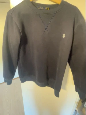 Mörkblå stickad tröja Polo Ralph Lauren - Mörkblå s tröja från Polo Ralph Lauren med klassisk rund halsringning och broderad vit logga på bröstet. Tröjan har ribbade muddar vid ärmslut och nederkant samt en avslappnad passform. Perfekt för en stilren och enkel look.