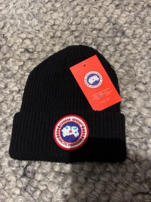 Svart ribbstickad mössa Canada Goose - Svart ribbstickad mössa från Canada Goose med klassisk uppvikt kant och det ikoniska runda Arctic Program-märket framtill. Perfekt för kalla vinterdagar och snygg till alla outfits. Materialet är mjukt och värmande.