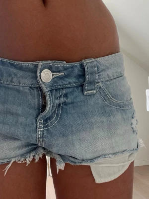 Lågmidjade jeansshorts i ljusblå denim - Säljer ett par riktigt snygga lågmidjade jeansshorts i ljusblå denim med slitna detaljer och fransig kant. Shortsen har klassiska fickor, bälteshällor och knappstängning framtill. Perfekta för varma sommardagar!