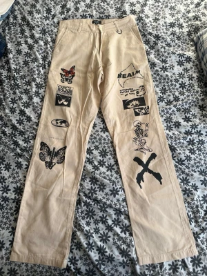 Beige chinos med tryck  - Unika beige chinos från bohoo med flera coola svarta tryck, bland annat fjärilar, skelett och text. Raka ben och klassisk passform. Perfekta för dig som vill sticka ut med din stil. Tillverkade i bomull för en skön känsla.