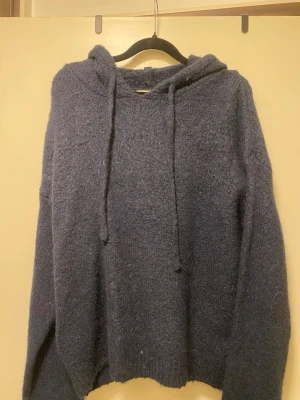 Mörkblå fluffig hoodie från Gina Tricot - Mysig mörkblå hoodie med huva och dragsko. Tröjan är stickad i ett fluffigt material som känns riktigt skönt mot huden. Perfekt för dig som gillar en avslappnad och chill stil. Långa ärmar och ribbade muddar ger en snygg passform.