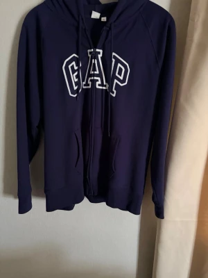 Mörkblå hoodie från GAP - Snygg mörkblå hoodie från GAP med vit broderad logga på bröstet. Klassisk modell med huva, dragsnören och känguruficka framtill. Perfekt för en avslappnad och trendig stil.