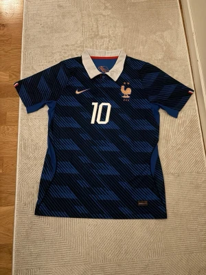 Frankrike fotbollströja Mbappé #10 - Snygg blå fotbollströja från Nike med Frankrikes landslagsmärke, nummer 10 och Mbappé på ryggen. Tröjan har korta ärmar, vit krage och diskret mönster i olika blå nyanser. Perfekt för fotbollsfans som vill visa sitt stöd för landslaget.
