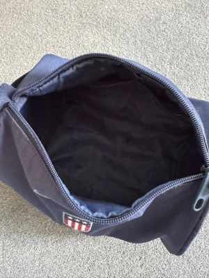 Marinblå necessär från GANT - Snygg marinblå necessär från GANT med dragkedja och broderad logotyp framtill. Tillverkad i slitstark textil och har ett praktiskt handtag på sidan. Perfekt för att organisera dina småsaker eller hygienartiklar på resan.(Pris kan presenteras)
