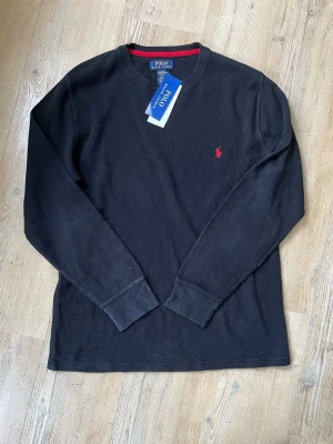 Svart långärmad tröja Polo Ralph Lauren - Svart långärmad tröja från Polo Ralph Lauren med rund halsringning och röd broderad logga på bröstet. Tröjan har en diskret röd detalj på insidan av kragen och är tillverkad i ett mjukt, strukturerat bomullstyg. Perfekt för en stilren och avslappnad look.