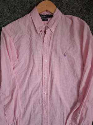 Rosa rutig skjorta från Ralph Lauren - Snygg rosa skjorta från Ralph Lauren med diskret rutigt mönster och klassisk knappstängning. Skjortan har lång ärm och ikonisk broderad logga på bröstet. Perfekt för dig som vill ha en stilren och fräsch look.