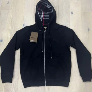 Svart Burberry hoodie med dragkedja - Svart hoodie från Burberry med klassiskt rutigt foder i huvan och snygga detaljer som silverfärgad dragkedja och snörning. Tröjan har långa ärmar, två fickor fram och ribbade muddar. Perfekt för dig som vill ha en stilren och lyxig look. Den är i väldigt bra skick då den bara testats någon enstaka gång