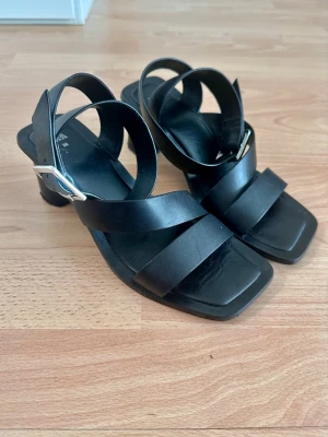 Svarta sandaler med klack och spänne - Stilrena svarta sandaler med breda remmar i konstläder och chunky klack. Snyggt fyrkantig tå och silverfärgat spänne på sidan. Perfekta för att lyfta din outfit med en modern touch.