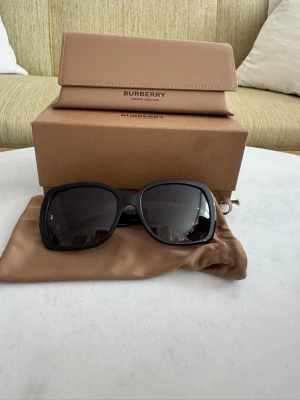 Burberry svarta solglasögon - Stilrena svarta solglasögon från Burberry med klassisk rektangulär form. Kommer med ett beige fodral och originalkartong. Perfekta för att lyfta din look med en touch av lyxig brittisk design. Äkta och aldrig använda.
