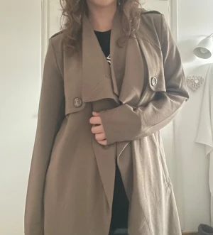 Brun kappa/trenchcoat - Snygg brun/beige trenchcoat med långa ärmar och knytbart bälte i midjan. Jackan har dekorativa knappar och axelklaffar. Materialet är lite tunnare så den passar bra på våren och hösten! Den är knappt använd och är från Nelly.