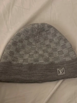  Grå Louis Vuitton mössa - Snygg grå mössa från Louis Vuitton med diskret rutmönster och broderad LV-logga framtill. Mjuk och stretchig passform, perfekt för att hålla värmen med stil. Passar dig som gillar exklusiva accessoarer och streetwear-vibe.