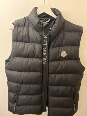 Svart dunväst från Moncler - Pris kan diskuteras! Säljer en stilren och varm Moncler dunväst i svart färg, storlek XL. Västen har den klassiska Moncler-loggan på bröstet, hög krage och två sidofickor med dragkedja. Perfekt för både vardag och höst/vinter.
