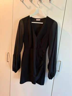 Svart klänning, Nelly - Säljer en svart kort klänning från Nelly. Klänningen har omlottdetalj, är v-ringad och har mesh ärmar som är lite genomskinliga. Klänningen har lite lätt rynkade detaljer i sidan och är tillverkad i ett mjukt och stretchigt material. 
