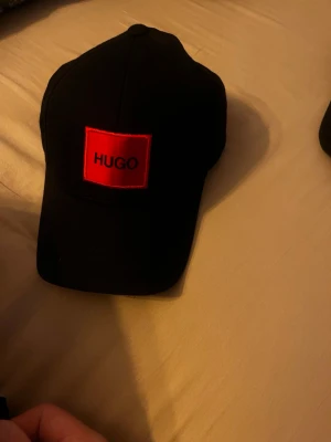 Svart keps från Hugo med röd logga - Snygg svart keps från Hugo med en tydlig röd patch framtill där det står HUGO. Kepsen har klassisk böjd skärm och justerbar rem baktill. Insidan har röda detaljer och etikett. Perfekt accessoar för en stilren look.