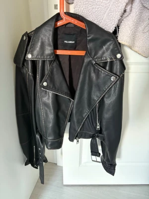 Svart skinnjacka från Pull&Bear - Snygg svart skinnjacka från Pull&Bear med klassisk bikerstil, silvriga dragkedjor och knappar samt bälte i midjan. Jackan har breda slag och coola detaljer som axelklaffar och fickor med dragkedja.