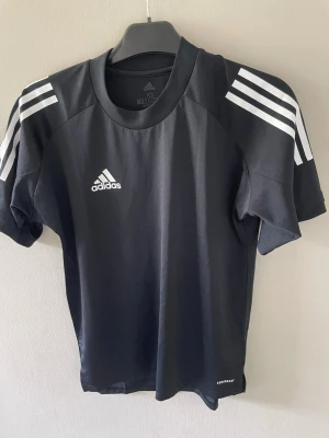 Svart Adidas sporttröja med vita ränder - Svart sporttröja från Adidas med klassiska vita ränder på axlarna och Adidas-logga på bröstet. Tillverkad i ett lätt och funktionellt material som andas, perfekt för träning eller sportaktiviteter. Rund halsringning och korta ärmar.