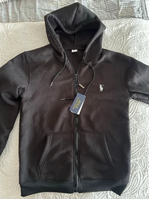 Svart hoodie från Polo Ralph Lauren - Svart hoodie med dragkedja från Polo Ralph Lauren. Klassisk design med huva och dragsko, två fickor framtill och broderad logga på bröstet. Mjuk bomullsblandning som är perfekt för en avslappnad stil.