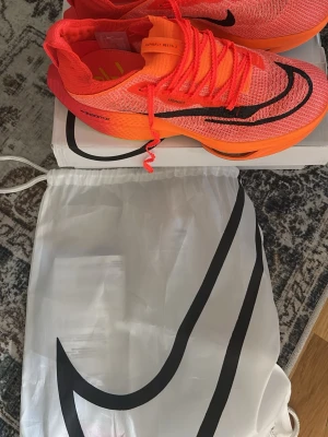 Nike löparskor - Säljer ett par Nike ZoomX Vaporfly Next% 2 i en riktigt snygg neonorange färg med svarta detaljer och limegröna inslag på sulan. Skorna har en lätt och ventilerande mesh-överdel, responsiv ZoomX-sula och klassisk Nike-logga. Perfekta för dig som vill maxa löpningen.