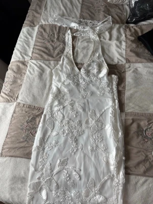 Vit spetsklänning med halterneck - Elegant vit långklänning med halterneck och öppen rygg. Klänningen har vackra spetsdetaljer och broderade blommor över hela tyget. Figurnära passform och transparenta partier ger en lyxig känsla. Perfekt för speciella tillfällen.