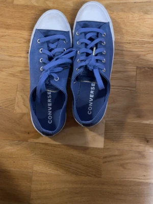 Blå Converse All Star låga sneakers - Snygga blå Converse All Star sneakers med vita gummisulor och tåhättor. Klassisk låg modell med textilöverdel, vita snören och metallöljetter. Perfekta för en avslappnad och cool stil.