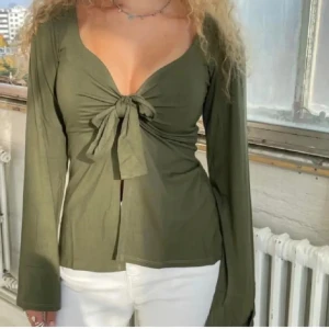 Olivgrön topp med knyt - Snygg olivgrön topp med v-ringning och knyt framtill. Långärmad design som ger en elegant look. Perfekt för att lyfta din outfit med stil. Sytt upp den lite vid bysten och hela magen, den är alltså inte öppen vid magen.