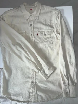 Vit jeansskjorta från Levi's - Säljer en stilren vit jeansskjorta från Levi's med klassisk krage och två bröstfickor med knappar. Skjortan har långa ärmar och silverfärgade tryckknappar. Perfekt för en avslappnad men snygg look.