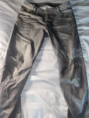 Gråa slim fit jeans W29xL32 - Snygga gråa slim fit jeans från G-RAW med klassisk femficksdesign och diskreta sömmar. Jeansen har en modern passform och är tillverkade i mjukt denimtyg som ger en skön känsla. Perfekta för dig som gillar stilrena och trendiga jeans.