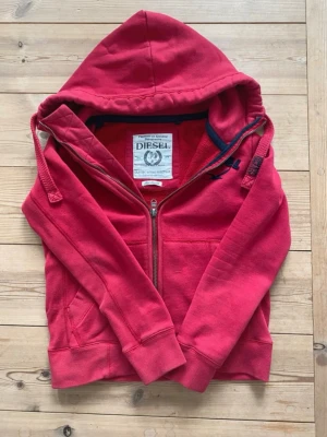 Diesel hoodie - Snygg röd hoodie från Diesel med dragkedja framtill och broderad logga i svart på bröstet. Hoodien har huva med dragsko, ribbade muddar och fickor på sidorna. Perfekt för en avslappnad och cool stil. Storlek S men sitter mer som XS.