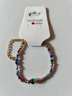 Färgglatt pärlarmband med gulddetaljer - Ett handgjort armband med små pärlor i olika färger som blått, rosa, silver och guld. Armbandet har en justerbar guldfärgad kedja och en större blå pärla i mitten. Perfekt accessoar för att ge din stil en färgklick.