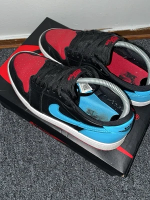 Nike Air Jordan 1 Low svart/röd/blå - Säljer ett par Nike Air Jordan 1 Low med svart ovandel, röd tåbox och blå detaljer på hälen och swoosh. Skorna har klassisk vit sula och svarta skosnören. Perfekt för dig som gillar sneakers med färg och ikonisk design.