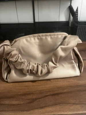 Beige kuvertväska med rynkad detalj - Stilren beige kuvertväska i satinliknande material med en unik rynkad axelrem. Väskan har dragkedja upptill och en mjuk, slät finish. Perfekt för dig som vill ha en trendig accessoar med det lilla extra.