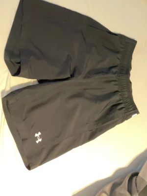 Svarta träningsshorts från Under Armour - Svarta träningsshorts från Under Armour med elastisk midja och logga på benet. Lätta och bekväma med en loose passform, perfekta för gymmet eller löpning. Klassisk design med vit logotyp och tryckt varumärke i midjan.