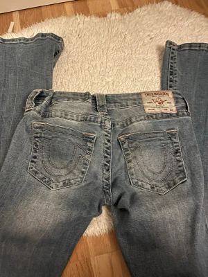 True Religion ljusblå bootcut jeans - Säljer ett par ljusblå bootcut jeans från True Religion med ikoniska bakfickor och kontrastsömmar. Jeansen har låg midja och klassisk femficksdesign. Perfekta för dig som gillar en avslappnad men trendig look.