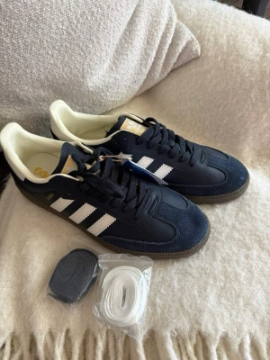 Adidas Samba OG mörkblå sneakers - Adidas Samba OG sneakers i mörkblå mocka och läder med vita detaljer och klassiska tre ränder på sidorna. Gummisula och vit hälkappa. Kommer med både  och vita skosnören. Perfekta för en stilren och sportig look.