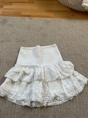 Vit volangkjol med broderade detaljer - Supersöt vit kortkjol med bred smock i midjan och två lager volanger. Kjolen har broderade spetsdetaljer och hålmönster som ger en romantisk känsla. Perfekt för varma dagar och lätt att matcha med en enkel topp.