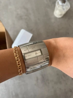 Silverfärgad bred klocka från DKNY - Stilren och bred klocka från DKNY i silverfärgat metallarmband. Klockan har en rektangulär urtavla med minimalistisk design och diskret DKNY-logga. Länkarna är blanka och ger ett modernt, chunky intryck. Perfekt accessoar för dig som gillar statement-smycken.