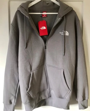 Grå hoodie från The North Face - Snygg grå hoodie från The North Face med dragkedja, huva och två stora fickor framtill. Diskret logga på bröstet och justerbara snören i huvan. Perfekt för en avslappnad och cool stil.
