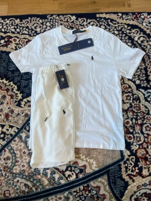 Vitt set från Polo Ralph Lauren - Snyggt vitt set från Polo Ralph Lauren med t-shirt och shorts. Båda plaggen har den klassiska broderade loggan på bröstet och benet. Setet är gjort i mjukt bomullsmaterial och har en avslappnad passform. Perfekt för en stilren och bekväm look.