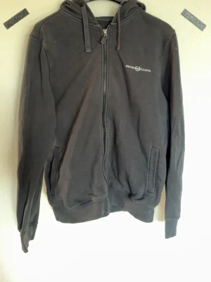 Svart hoodie från Henri Lloyd - Svart zip-hoodie från Henri Lloyd med dragsko i huvan och två fickor framtill. Klassisk design med logga på bröstet och ribbade muddar. Perfekt för en avslappnad och stilren look.