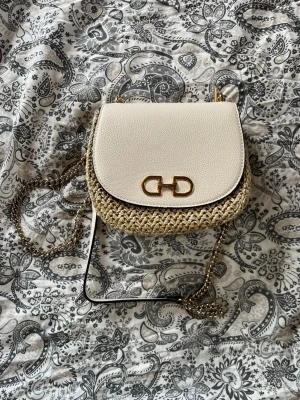 Beige axelremsväska med kedja - Snygg handväska i beige med flätad botten och lock i läderimitation. Väskan har guldfärgad kedjerem och dekorativt metallspänne framtill. Perfekt för dig som gillar trendiga accessoarer med en mix av material.