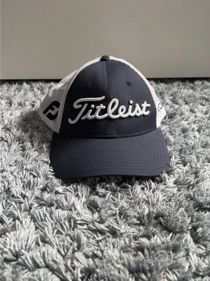 Titleist keps i navy och vitt - Snygg keps från Titleist i navyblått med vita meshpaneler och broderad logga framtill. På sidan står det 'Pro V1' och kepsen har böjd skärm. Perfekt för golfbanan eller en sportig look. Materialet är en kombination av bomull och mesh.