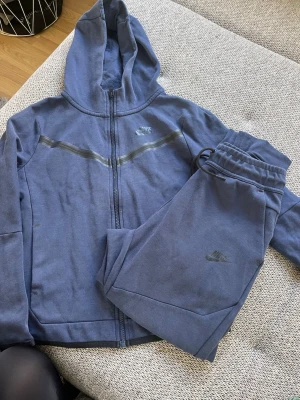 Blå Nike loungewear set med huva - Set från Nike med ziptröja och matchande byxor. Tröjan har huva och dragkedja samt diskreta reflexdetaljer över bröstet. Byxorna har elastisk midja och Nike-logga på benet. Bra skick, förutom ett litet hål på hoodien, se bild. 